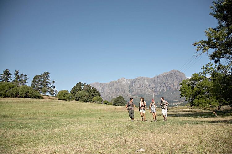 Gruppe junger Menschen mit Rucksäcken wandert über eine Wiese, mit Bergen im Hintergrund.