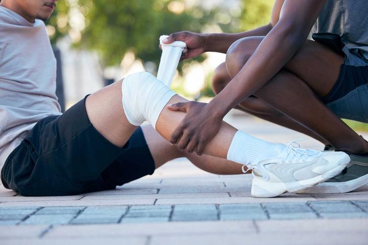 Bildnachweis: © stock.adobe.com / Talia M/peopleimages.com Sportler sitzt auf dem Boden und bekommt einen Verband am Knie