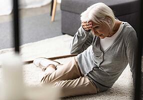 Seniorin sitzt am Boden und hält sich die Hand an die Stirn