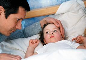 Kleines Mädchen liegt krank im Bett, der Vater legt ihr liebvevoll die Hand auf den Kopf.