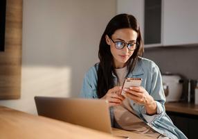 Frau schaut auf ihr Handy während sie am Laptop arbeitet