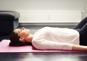 Junge Frau liegt mit geschlossenen Augen und angezogenen Beinen auf einer Yogamatte.