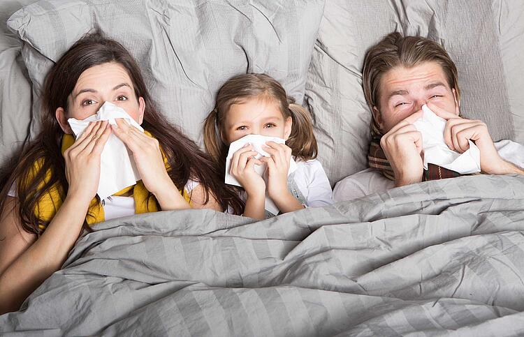 Hausmittel bei Schnupfen: Familie liegt krank im Bett und putzt sich die Nase