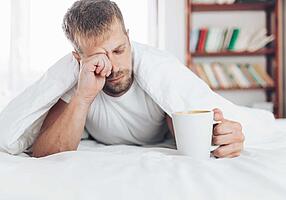 Verschlafen wirkender Mann trinkt im Bett eine Tasse Kaffee