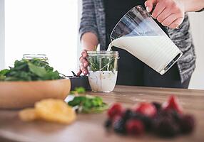 Eine Hand gießt Milch aus einem Krug in ein Glas mit Kräutern.