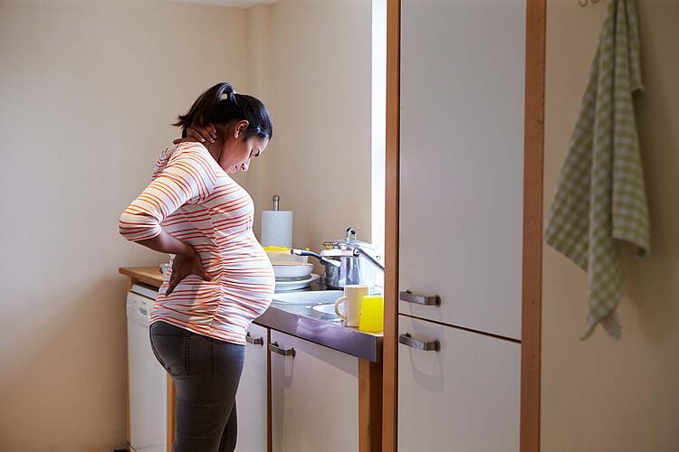 Hochschwangere Frau steht in der Küche vor der Spüle und hält sich den schmerzenden Rücken.
