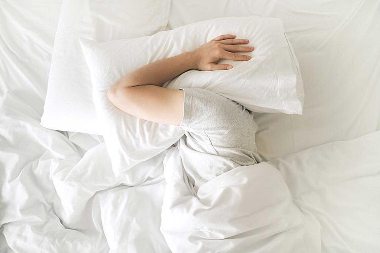 Müde Person hält sich ein Kopfkissen über den Kopf um weiter zu schlafen
