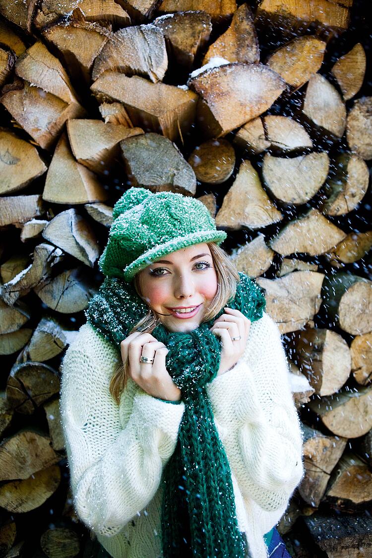 Frau mit Mütze und Schal steht vor einem Holzstapel und ist voller Schneeflocken.