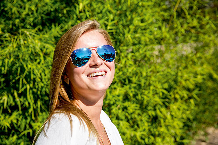 Junge Frau mit verspiegelter Sonnenbrille vor einer grünen Hecke.
