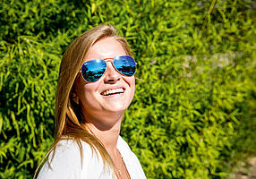 Junge Frau mit verspiegelter Sonnenbrille vor einer grünen Hecke.