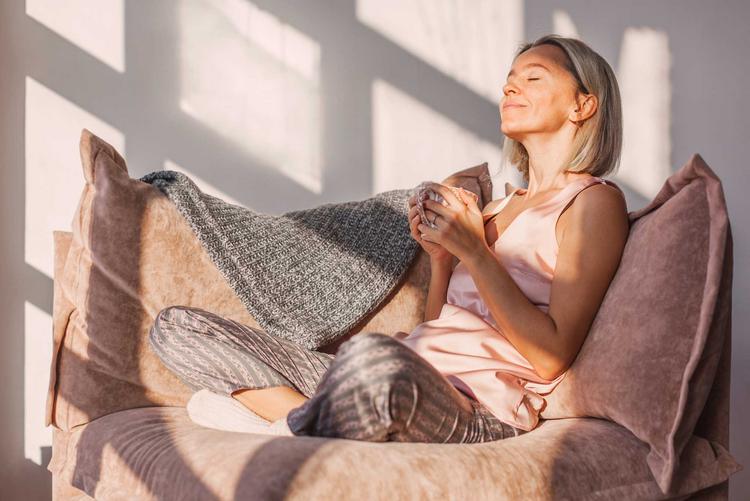 Eine Frau entspannt bei einer Tasse Tee auf dem Sofa, die Sonne scheint ihr ins Gesicht