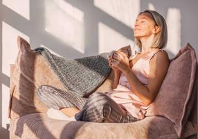 Eine Frau entspannt bei einer Tasse Tee auf dem Sofa, die Sonne scheint ihr ins Gesicht