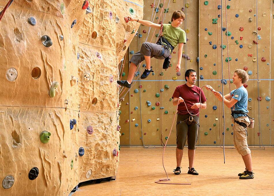 Drei junge Männer in einer Kletterhalle, zwei stehen am Boden, einer klettert an einer Wand.