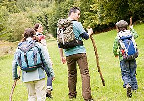 Familie mit Rucksäcken und Wanderstöcken wandert üebr eine Wiese auf einen Wald zu.