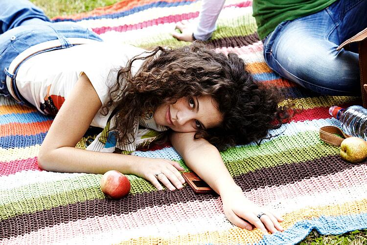 Junge Frau liegt entspannt auf einer Decke, einen Apfel und ein Handy vor sich.