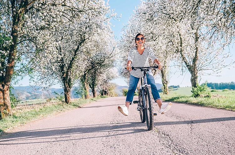 Frühlingsgefühle: Frau fährt mit dem Fahrrad eine Straße mit blühenden Bäumen entlang