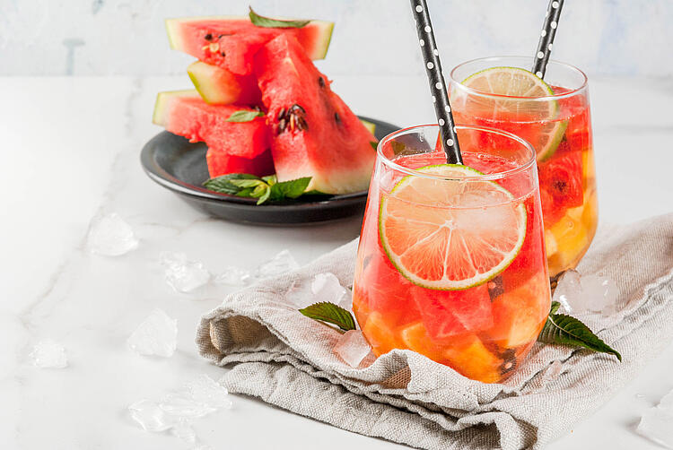 Zwei Gläser Melonen-Bowle mit Limettenscheiben und Strohhalmen, dahinter ein Teller mit Wassermelonen-Stücken.